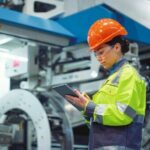 Woman_supervisor_looking_tablet_computer_in_huge_manufacturing_company._Focused_industrial_worker_analyzing_project_modern_plant._Safety_uniform_engineer_check_business_on_digital_tad_at_factory.