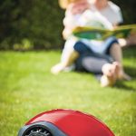 Close-up_of_a_modern_autonomous_mower_on_green_grass_in_front_of_a_young_mother_and_her_child_in_a_sunny_day
